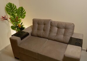 A beige sofa with a built-in stove, positioned against a plain wall with a large green plant. - Hotel Pousada Elo Inn (Jaboatão dos Guararapes)