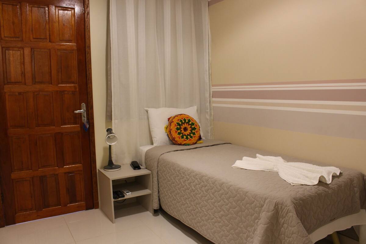 A bedroom with a wooden door, a bedside table with a lamp, a bed with a quilted bedspread, and a pillow with a colorful design.