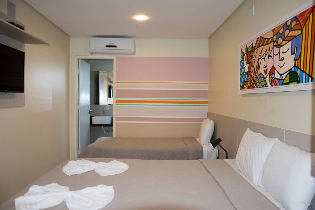 A hotel room with two beds, a TV, a painting, and a bathroom visible through an open door.