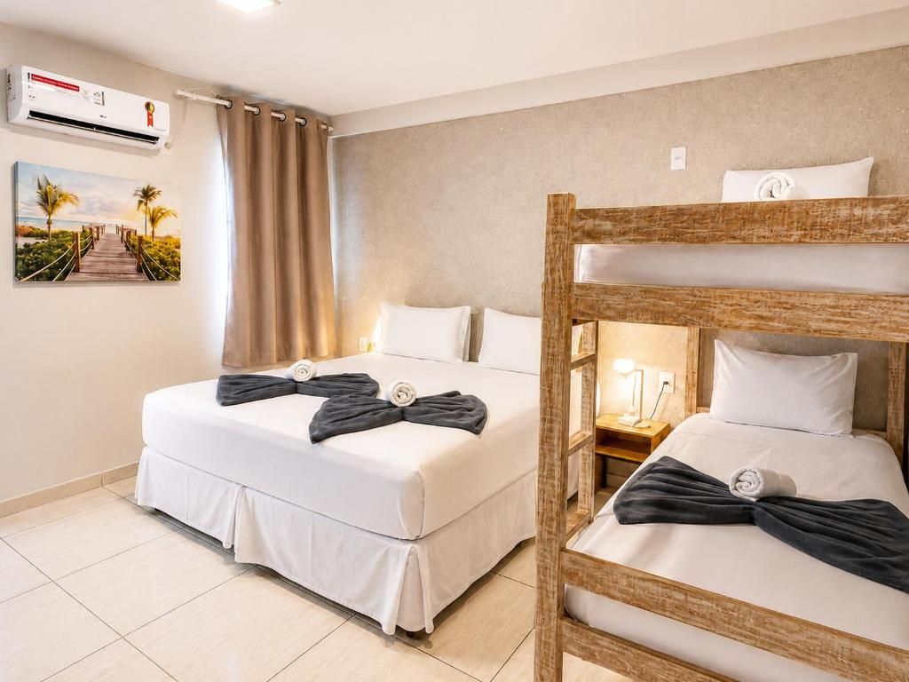 Quarto de hotel com duas camas, uma cama beliche de madeira, ar-condicionado fixado na parede, uma pintura de uma praia e uma cortina.