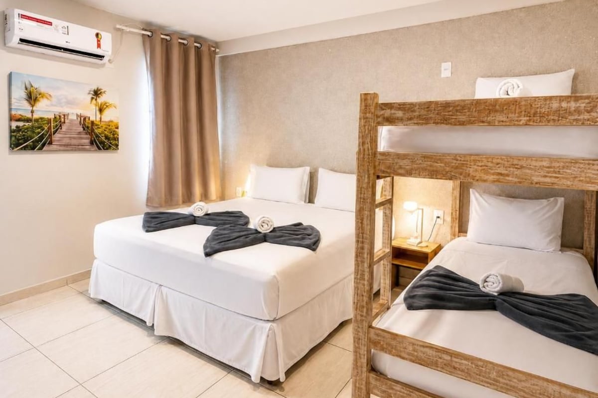 A hotel room with two beds, a wooden bunk bed, a wall-mounted air conditioner, a painting of a beach, and a curtain.