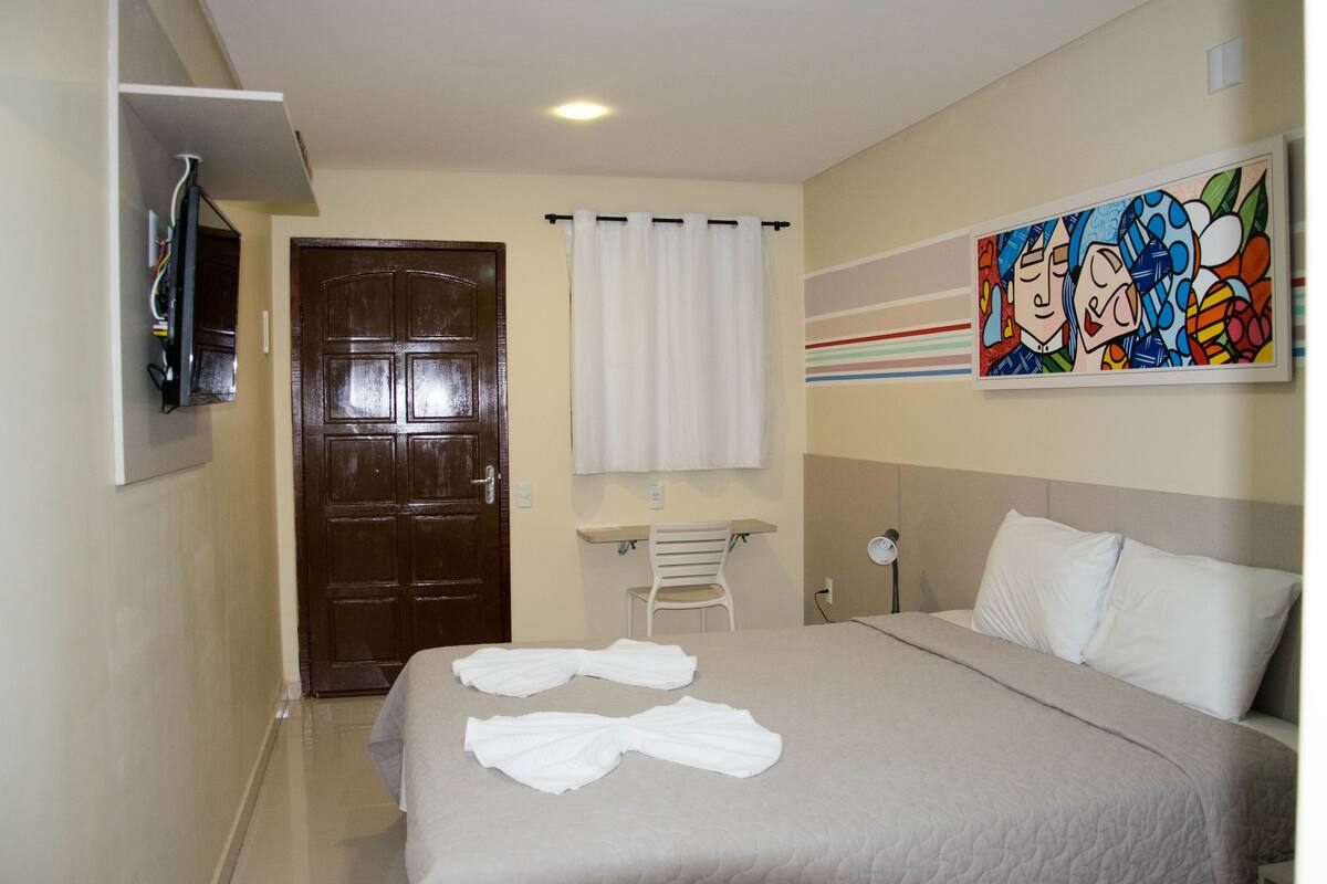 A hotel room with a bed, a desk, a chair, a TV mounted on the wall, and a painting above the bed.