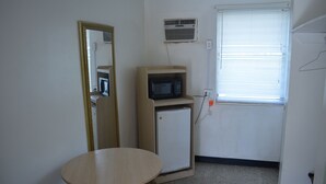 Una habitación pequeña con una mesa de madera, un microondas, un refrigerador pequeño, un espejo y una ventana con persianas.
