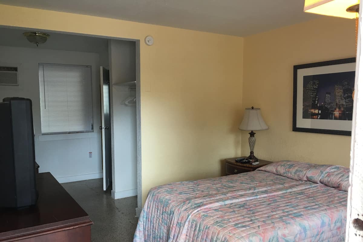 A hotel room with a bed, a nightstand with a lamp, a television, and a door leading to another room.