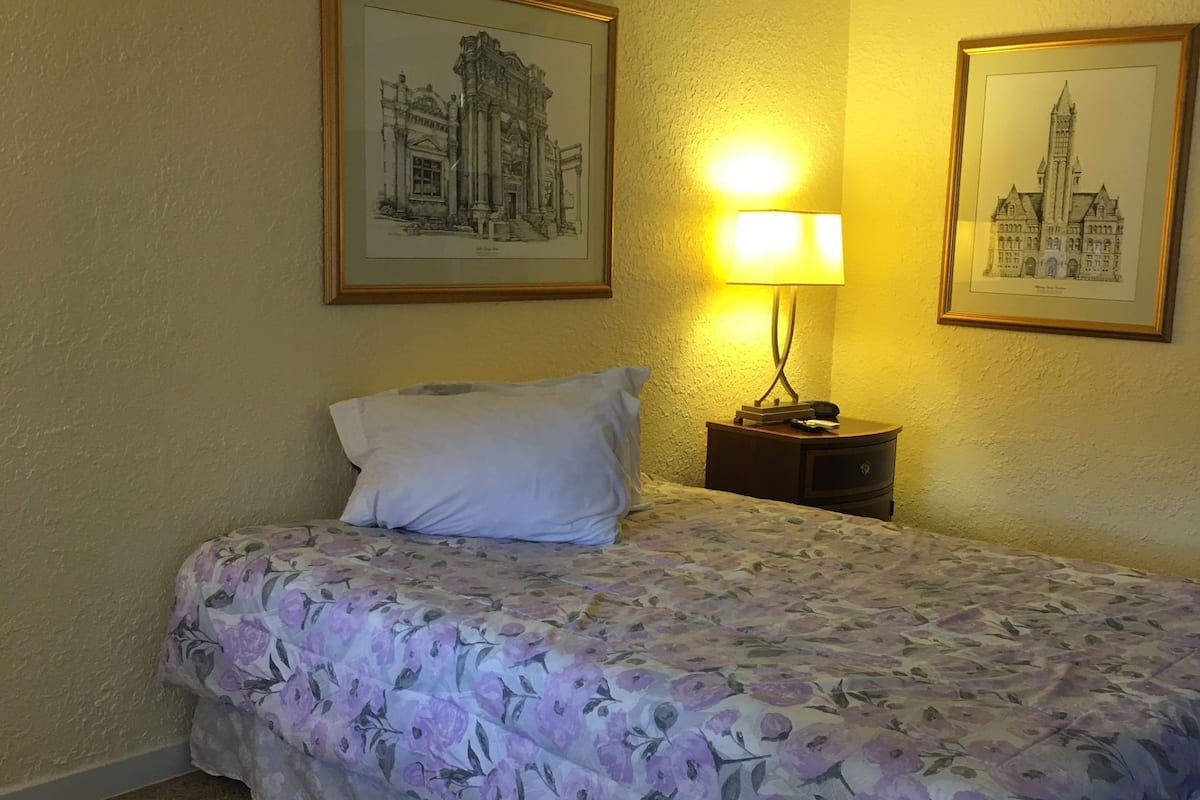 A hotel room with a bed, a nightstand, a lamp, and two framed pictures on the wall.