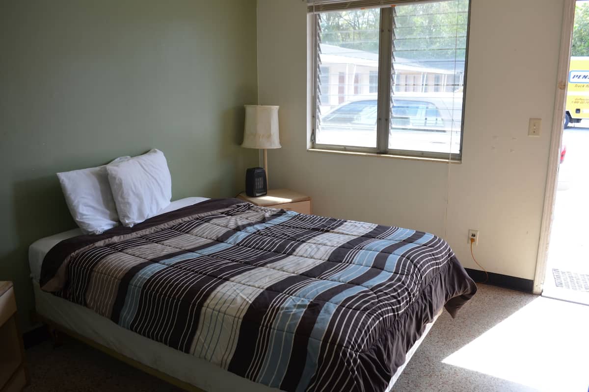 A single bed with a striped bedspread, a bedside lamp, and a window with a view of a parking lot.