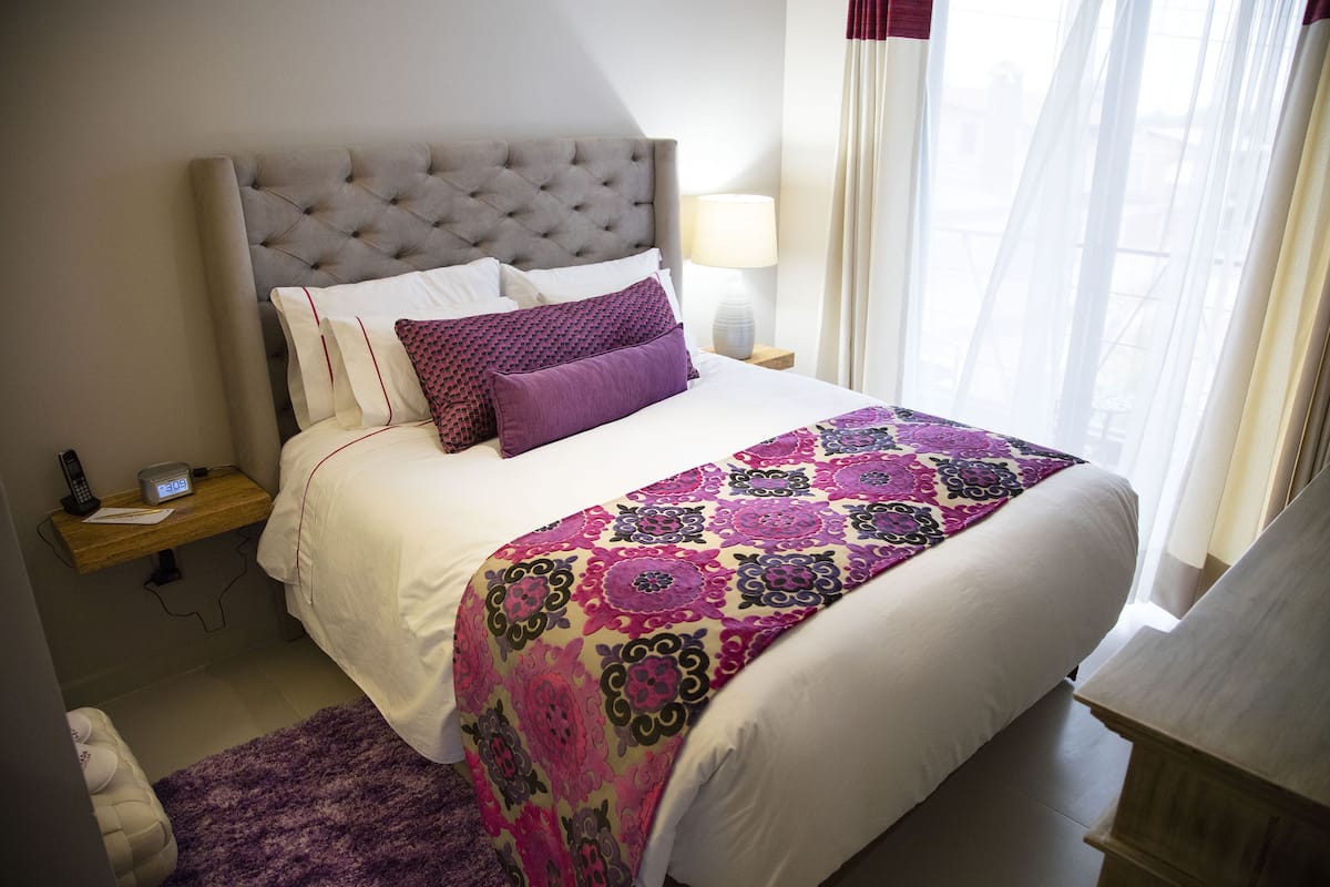 A neatly made bed with a patterned bedspread, a bedside table with a lamp, and a window with sheer curtains.