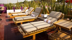A rooftop lounge area with wooden loungers, beige cushions, and a wooden table.