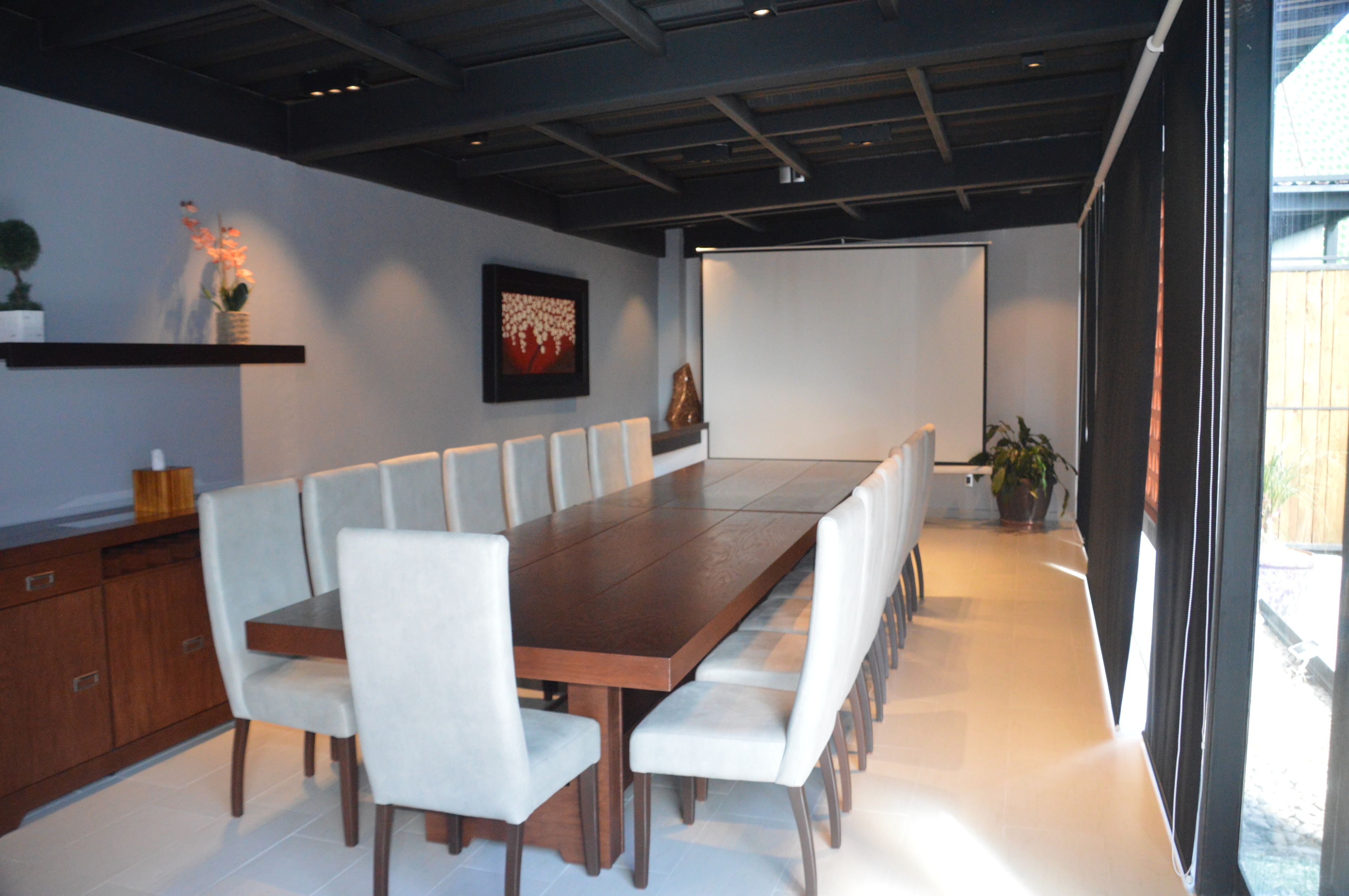 A modern conference room with a long wooden table, white chairs, and a potted plant.