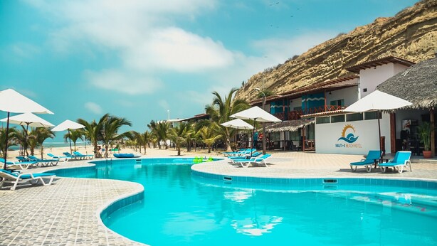 Una piscina con sillas de descanso y sombrillas, un edificio de techo de paja y palmeras.