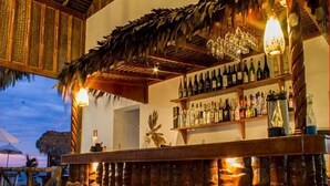 Zona de bar con temática tropical, con techo de paja, estantes de madera con botellas y una palmera decorativa.