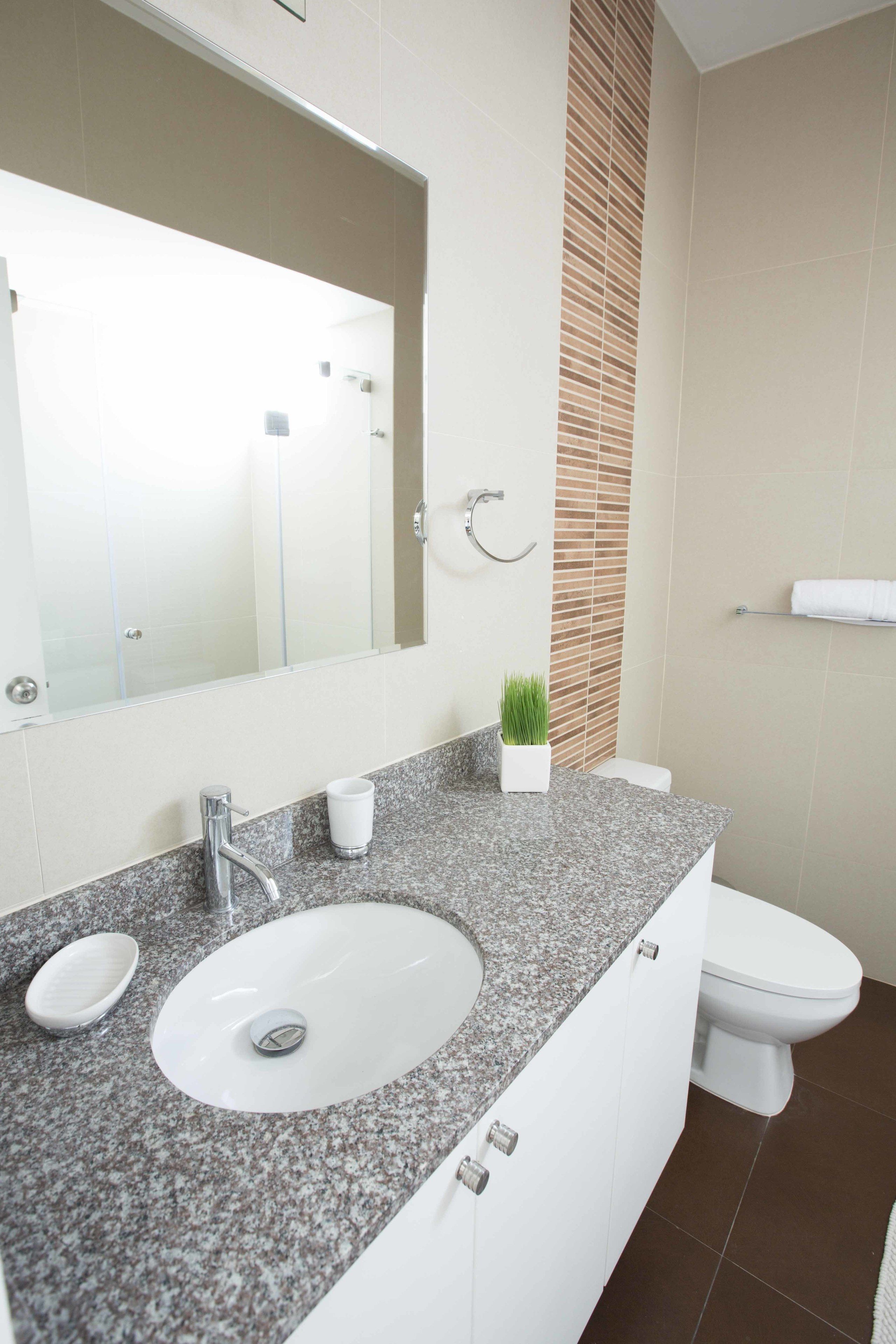 A modern bathroom with a granite countertop sink, a mirror, a toilet, and a towel rack.