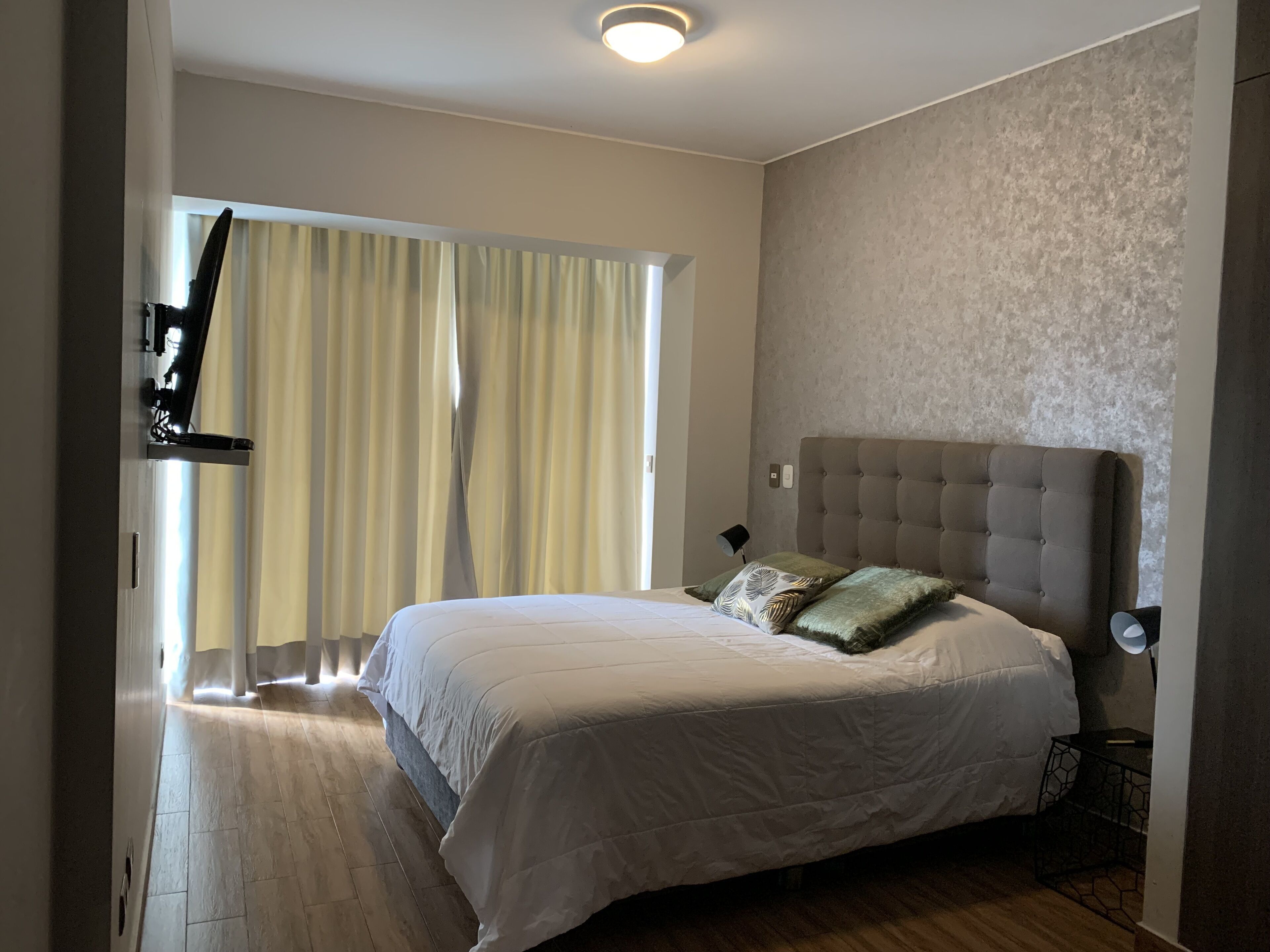 Un dormitorio con una cama grande, una ventana con cortinas, un televisor montado en la pared y una mesita de noche con lámpara.