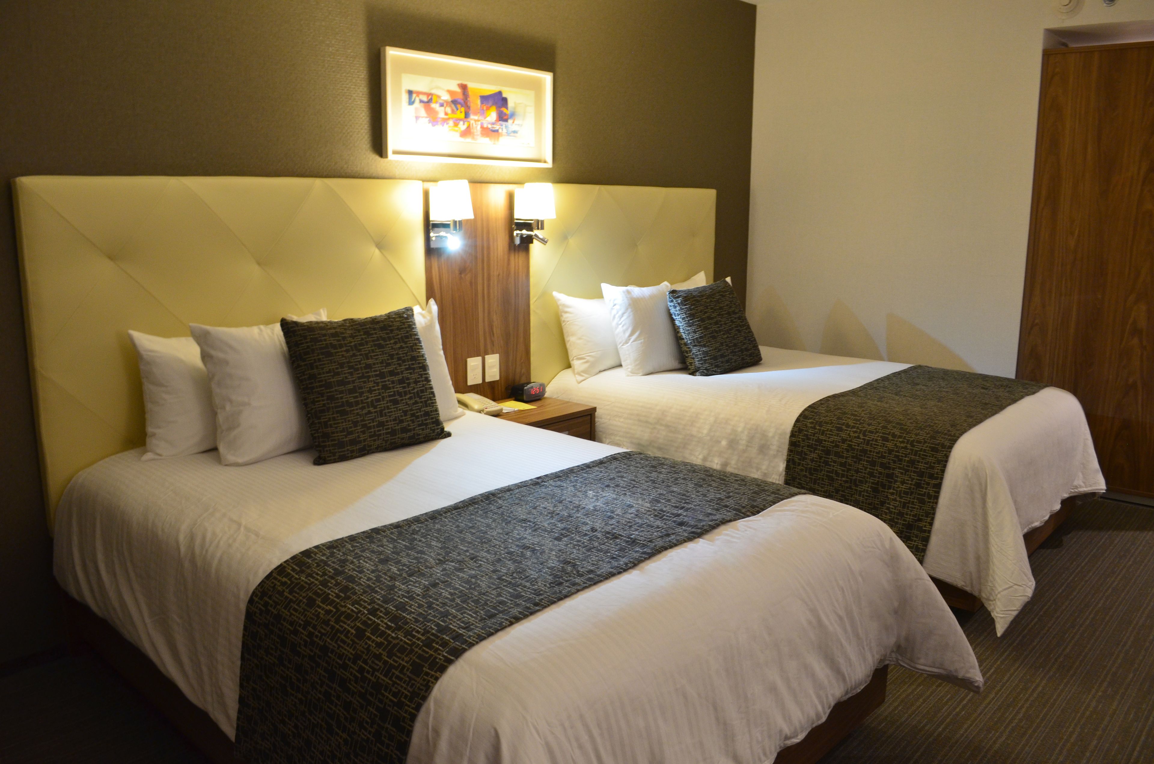 Hotelkamer met twee bedden, elk met witte lakens en donkere plaids. Houten hoofdeinde, nachtkastje met lamp en een lijst met een afbeelding boven de bedden.