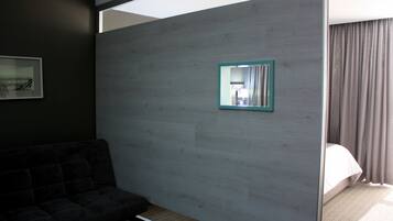 A modern hotel room with a dark-colored sofa, a wooden partition, a small mirror, and a bed with curtains.