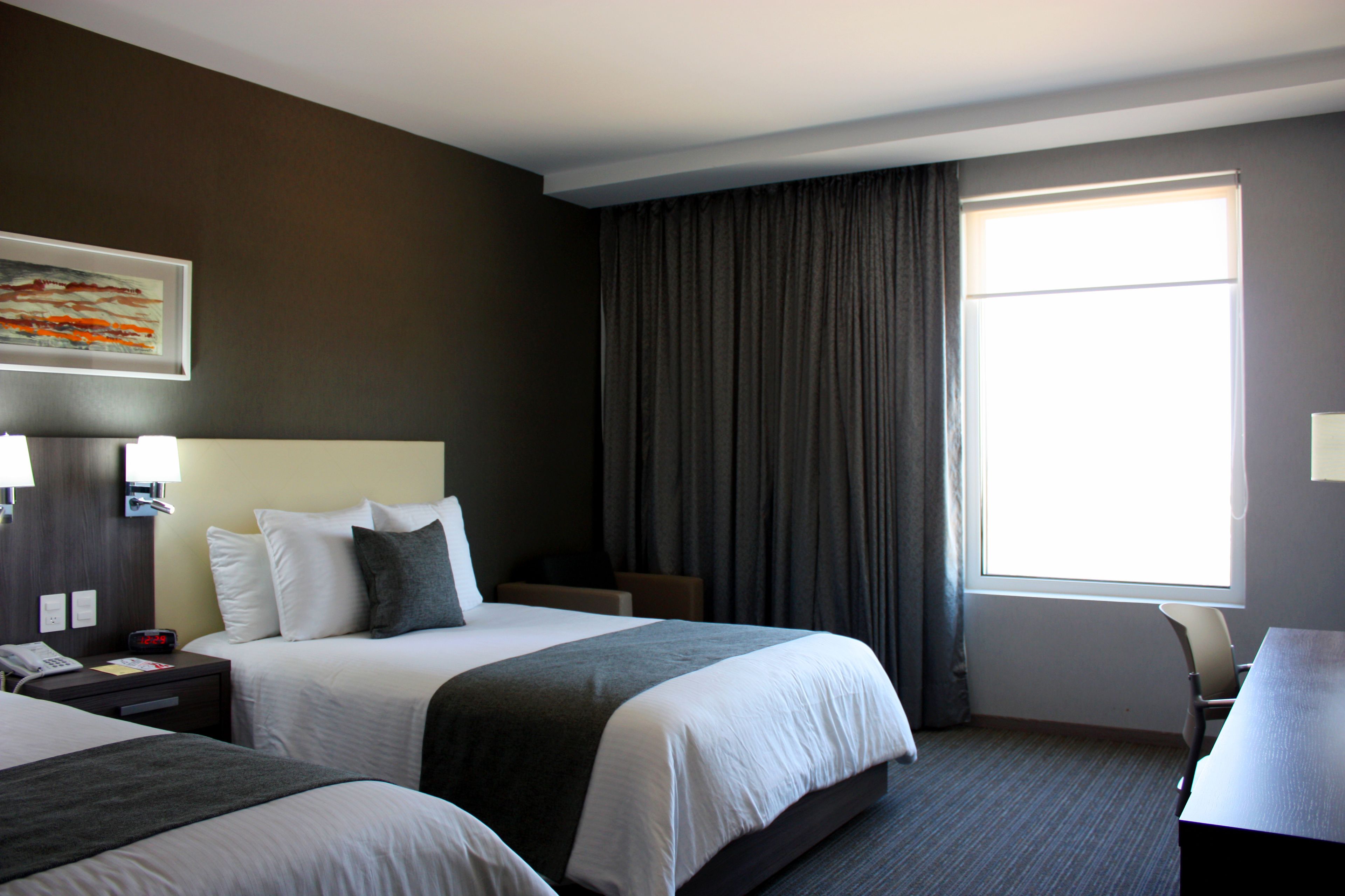 Quarto de hotel com duas camas, uma mesa, uma cadeira, uma mesinha de cabeceira com telefone e uma janela com cortinas.