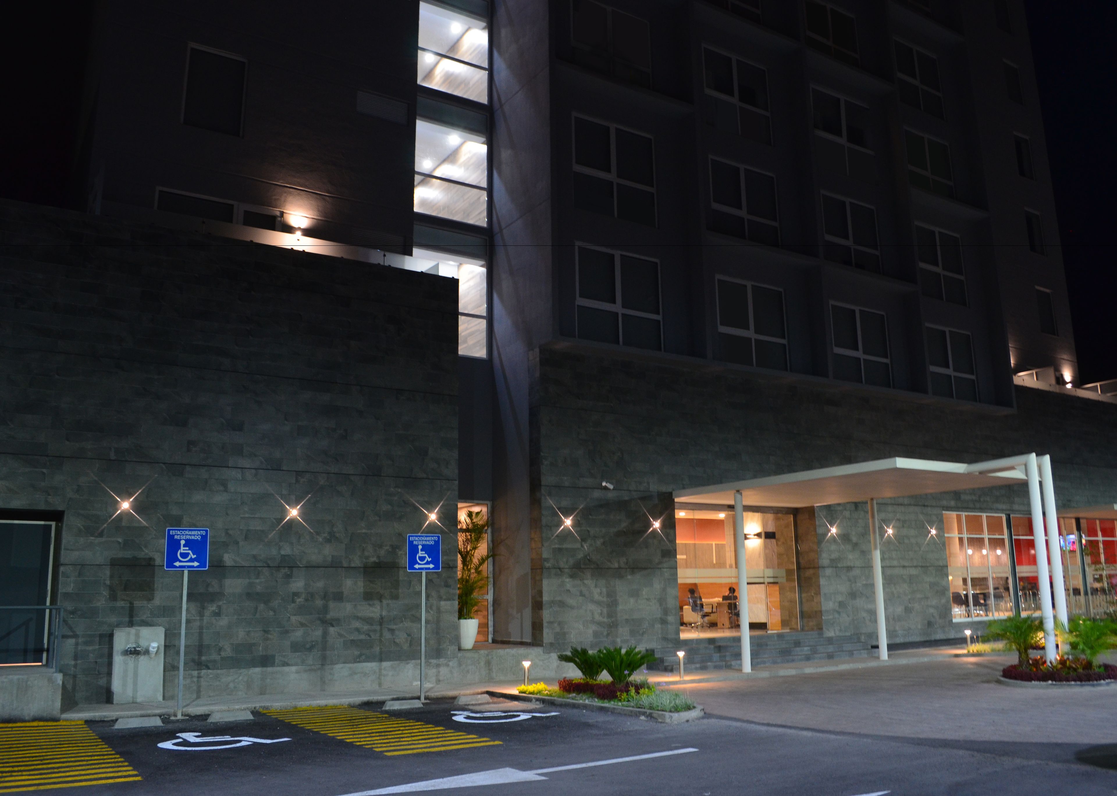 Un edificio moderno con un área de estacionamiento señalada para acceso de personas con discapacidad, una entrada cubierta y una fachada bien iluminada durante la noche.