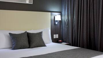 Een net opgemaakt bed met wit en grijs beddengoed, een nachtkastje met een lamp, en een donker gordijn.
