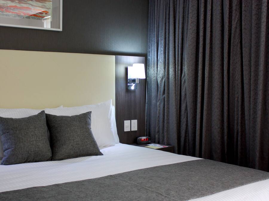 Een net opgemaakt bed met wit en grijs beddengoed, een nachtkastje met een lamp, en een donker gordijn.
