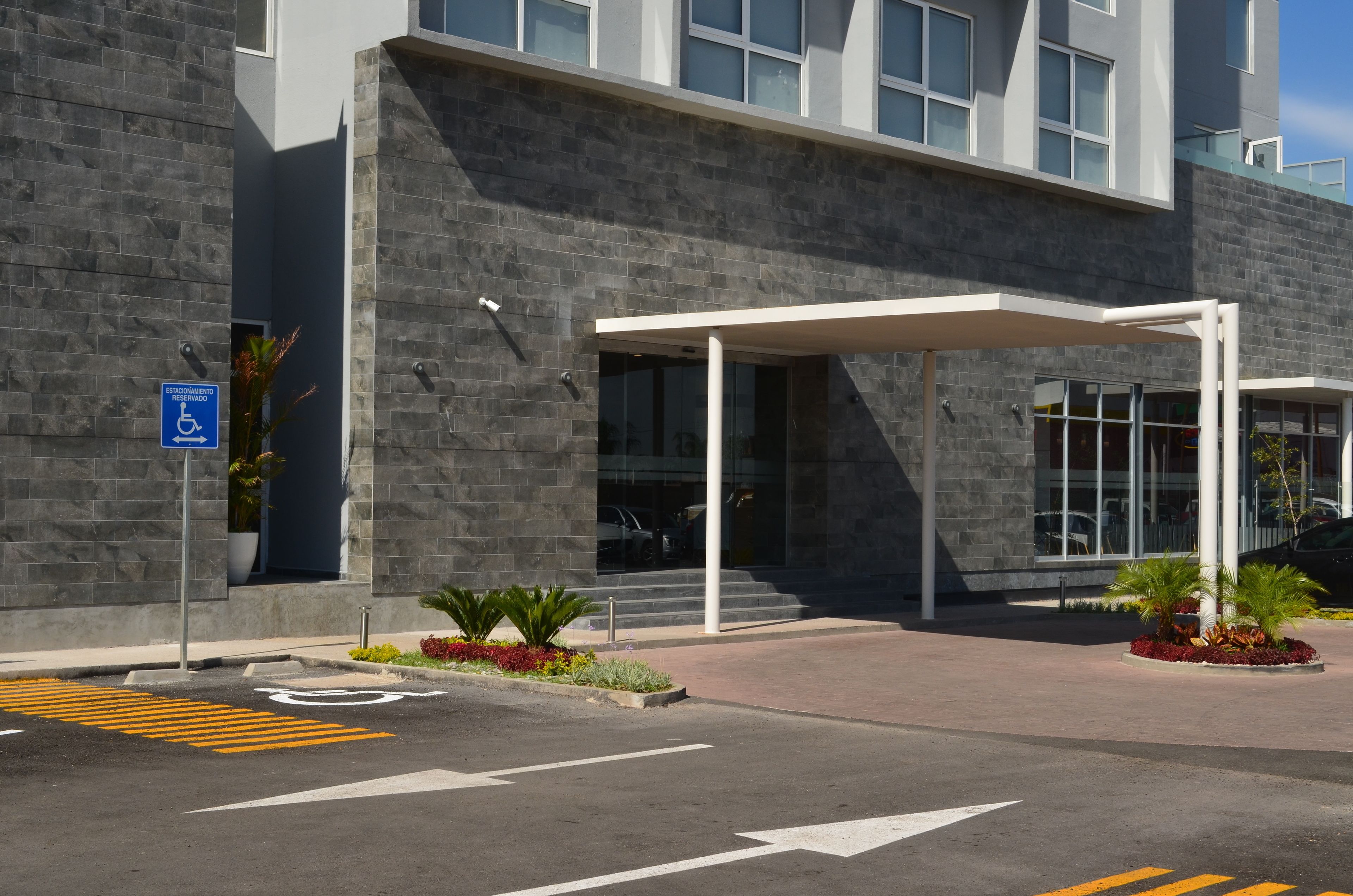 A modern building entrance with a covered walkway, a disabled parking sign, and a landscaped area with plants.