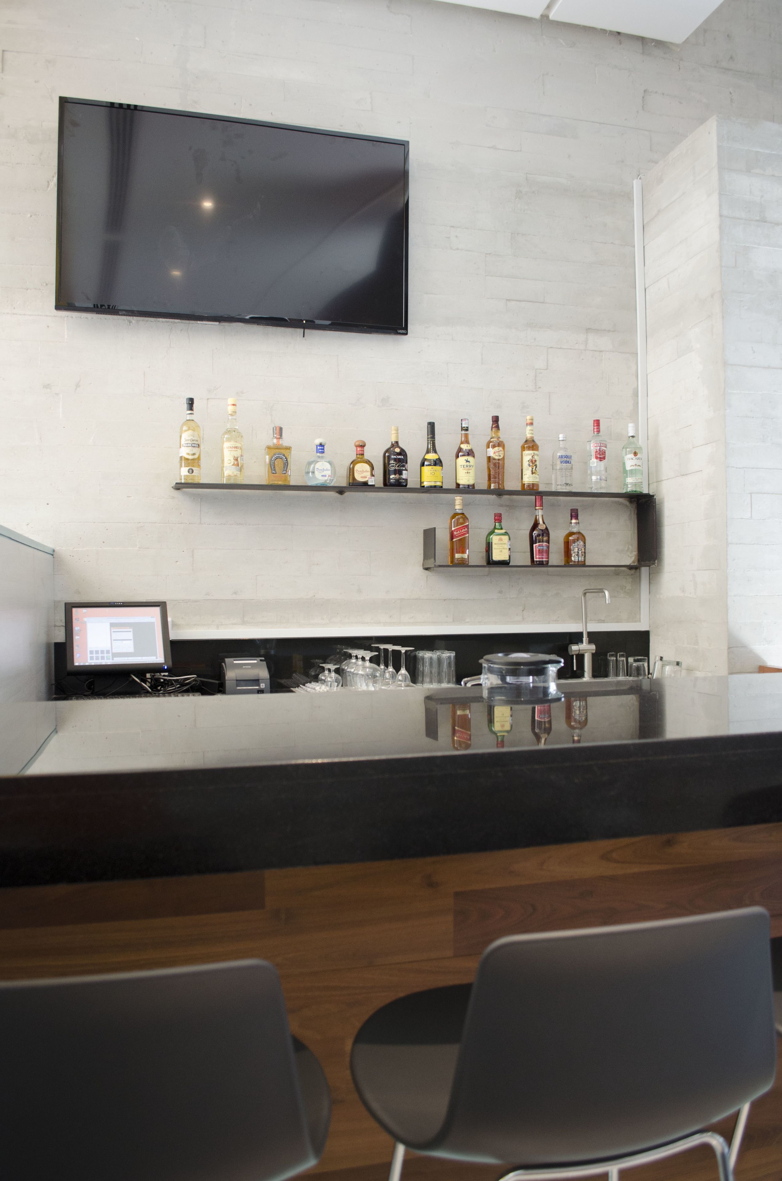 Un espace bar moderne doté d’un téléviseur à écran plat fixé au mur, de plusieurs étagères remplies de bouteilles d’alcool, d’un comptoir avec un évier et de deux chaises.