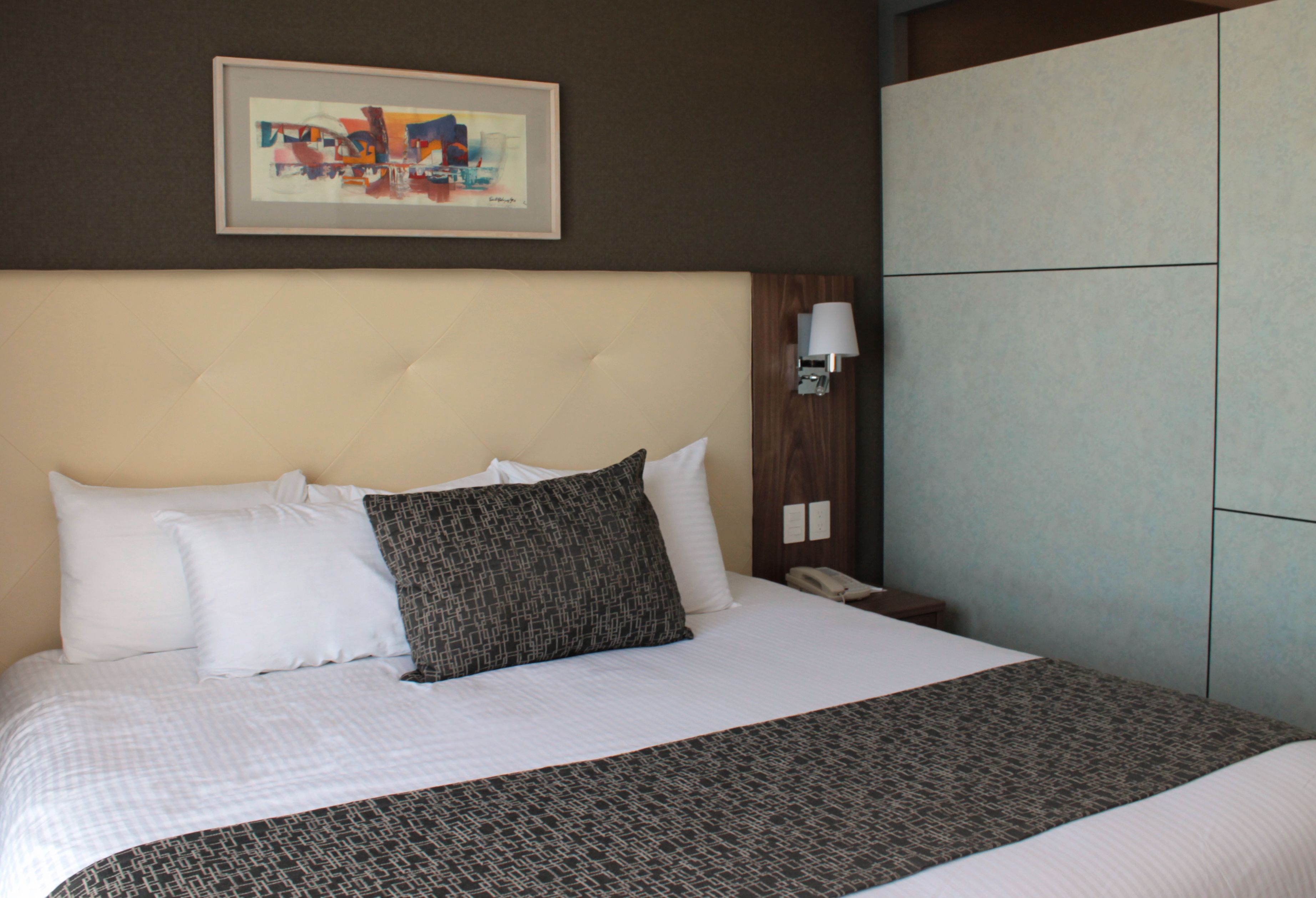 A hotel room with a large bed, a headboard, a bedside table with a lamp, and a framed artwork on the wall.