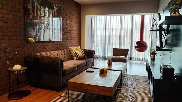 Una sala moderna con una pared de ladrillo, un ventanal con cortinas translúcidas, un sofá marrón, una mesa de centro de madera y un globo rojo colgando.