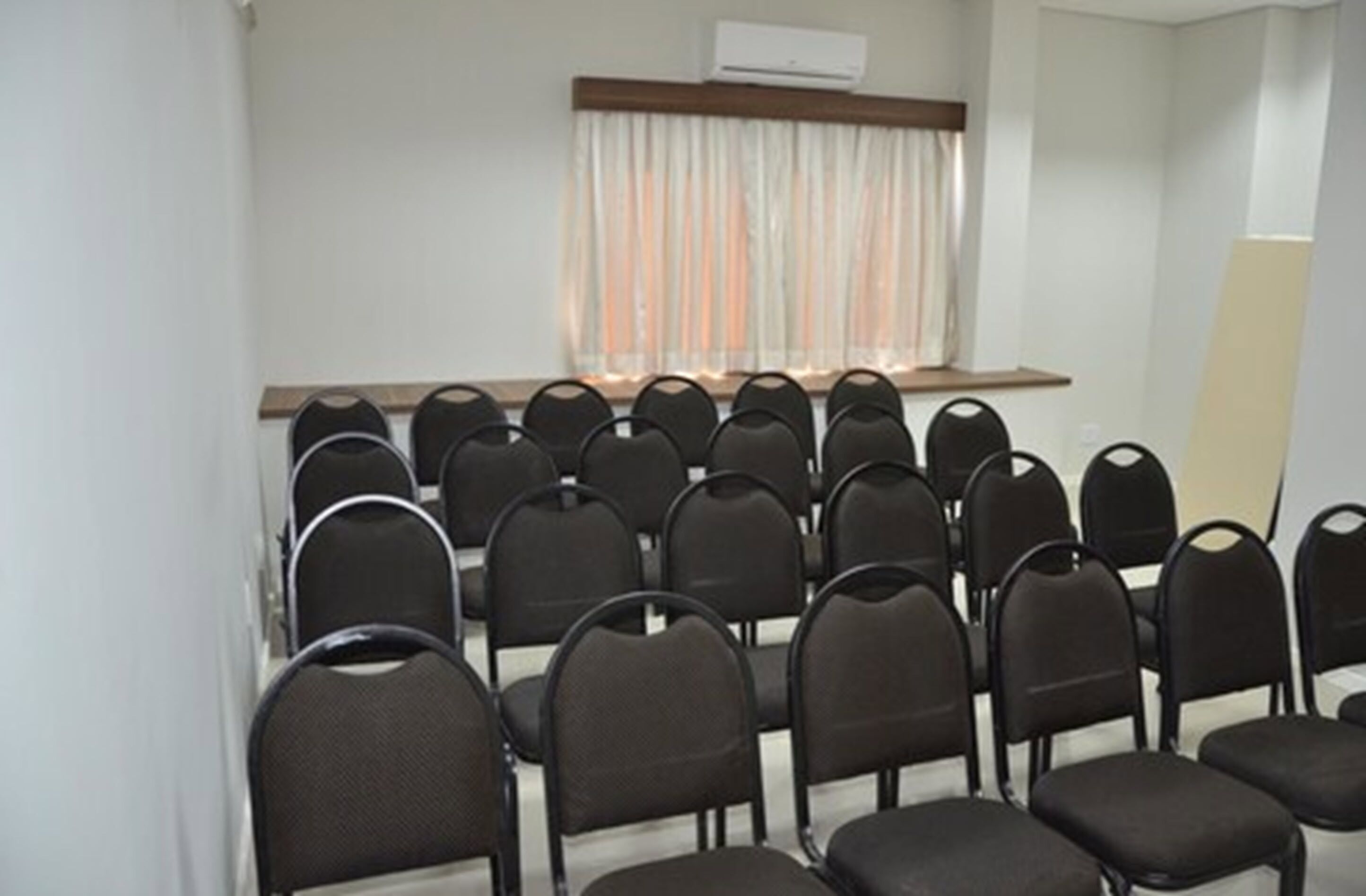 Un salón de conferencias con filas de sillas negras, una mesa de madera y un ventanal con cortinas.