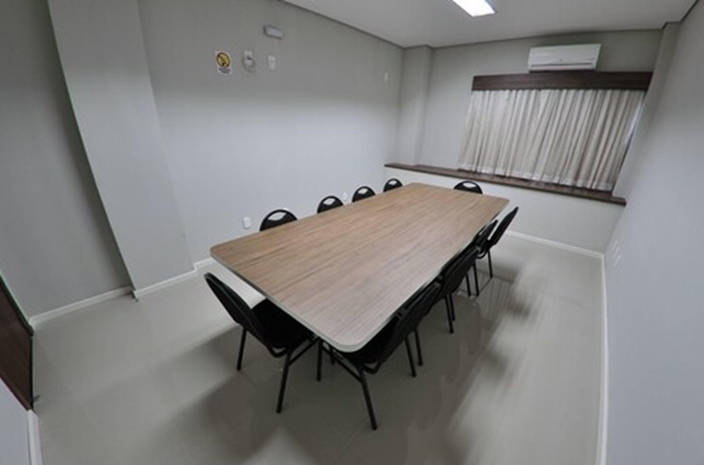 A conference room with a long table and chairs, a window with curtains, and an air conditioning unit on the wall.