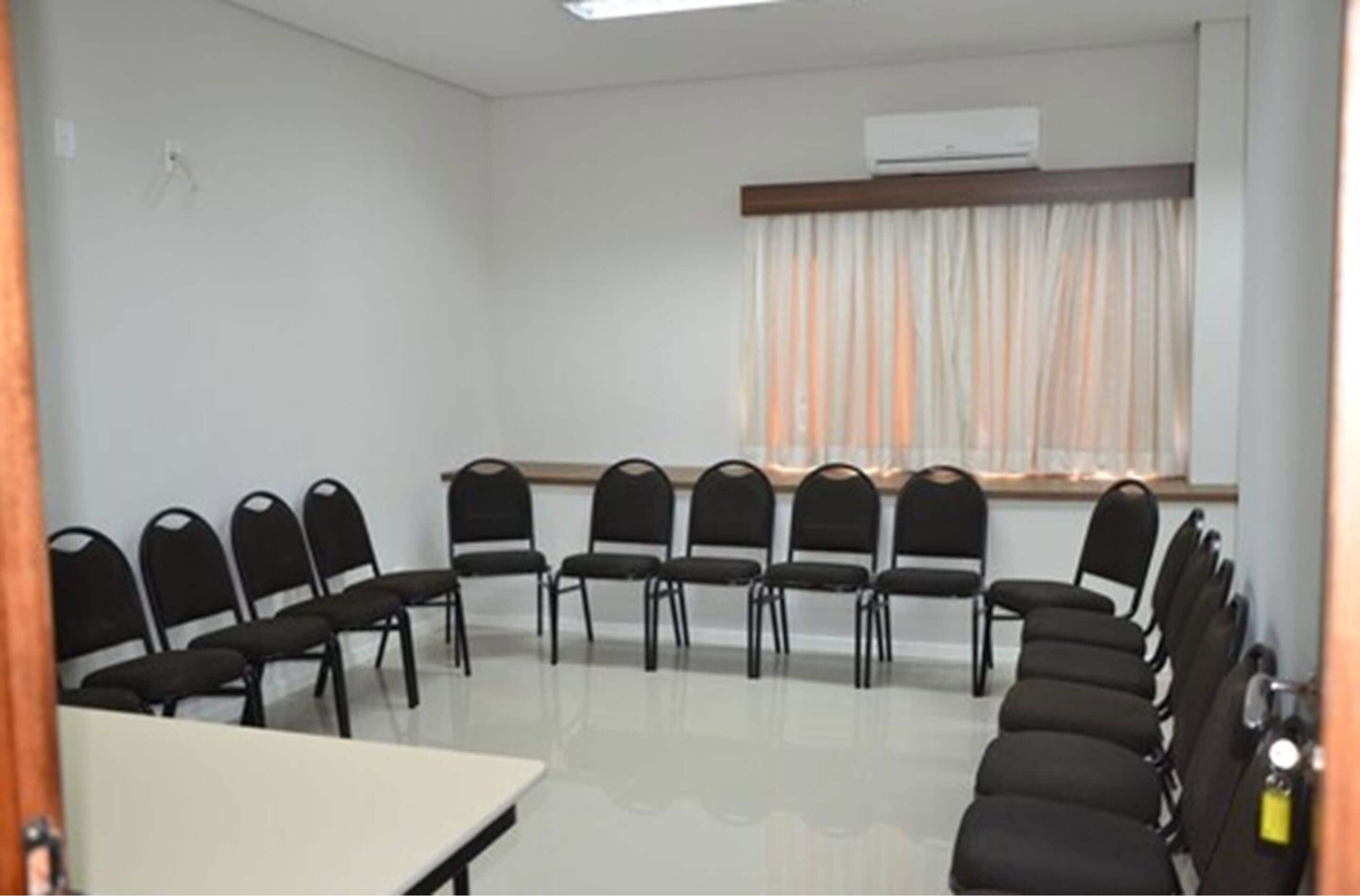 Un salón de conferencias con una mesa blanca, sillas negras, una ventana con cortinas y un equipo de aire acondicionado.