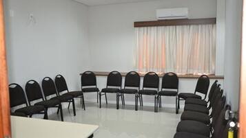 Un salón de conferencias con una mesa blanca, sillas negras, una ventana con cortinas y un equipo de aire acondicionado.