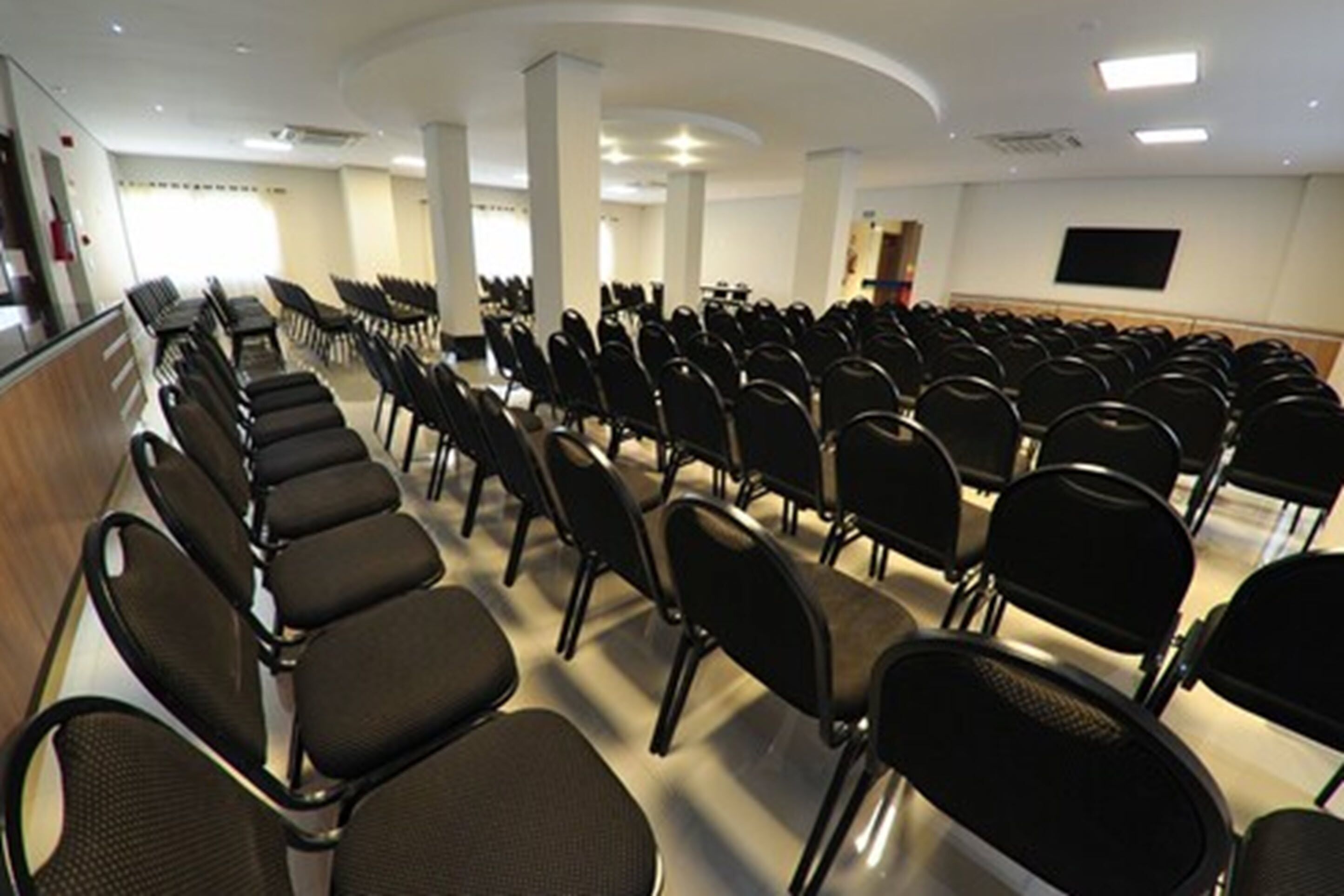 Une salle de conférence avec des rangées de chaises noires, un plafond blanc et un téléviseur à écran plat fixé au mur.