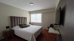 A bedroom with a bed, bedside table, lamp, window, and a flat-screen TV mounted on the wall. - MLA apartments - Alcanfores (Lima)