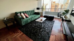 A living room with a green sofa, a coffee table, a black rug, and a view of buildings through large windows. - MLA apartments - Alcanfores (Lima)