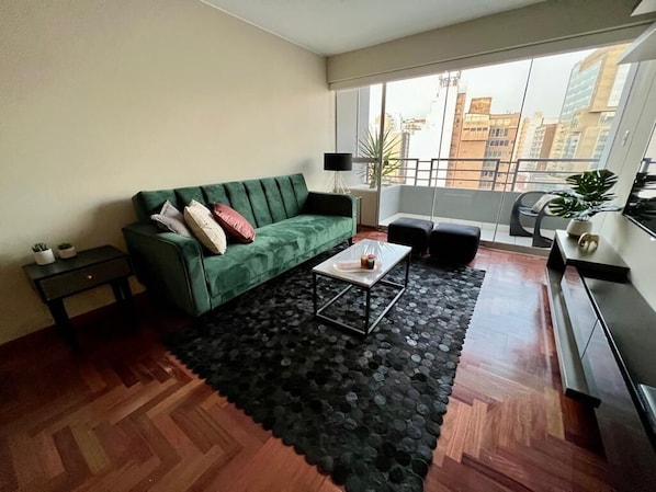 Een woonkamer met een groene bank, een klein bijzettafeltje, een zwart tapijt en uitzicht op de stad door grote ramen.