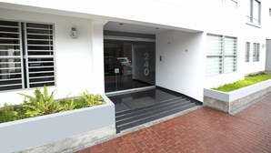 A modern building entrance with a glass door, a number '240' sign, and a brick pathway leading to the entrance. - MLA apartments - Alcanfores (Lima)