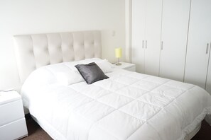 Un lit bien fait, avec une couette blanche et un seul oreiller noir, une lampe de chevet et une table de chevet blanche.