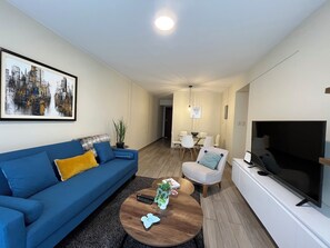 Un salon moderne avec un canapé bleu, une table basse, un téléviseur et, au fond, un coin repas.