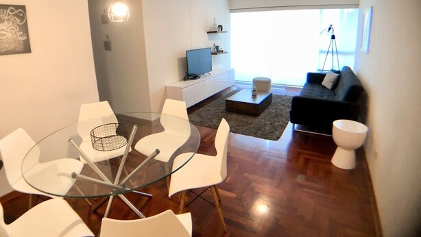 Ein modernes Wohnzimmer mit einem Glastisch, weißen Stühlen, einem schwarzen Sofa und einem Fernseher auf einer weißen Konsole.