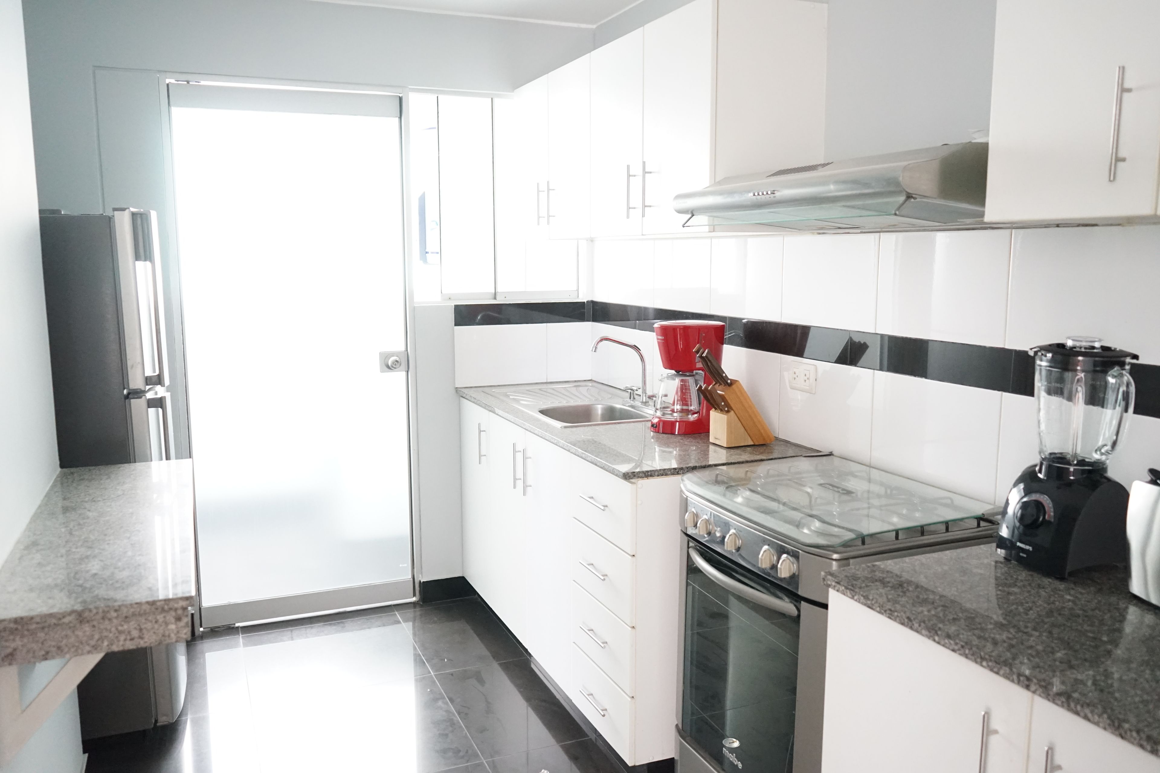 Una cocina moderna con armarios blancos, un refrigerador de acero inoxidable, una estufa a gas, un fregadero y una licuadora sobre la encimera.