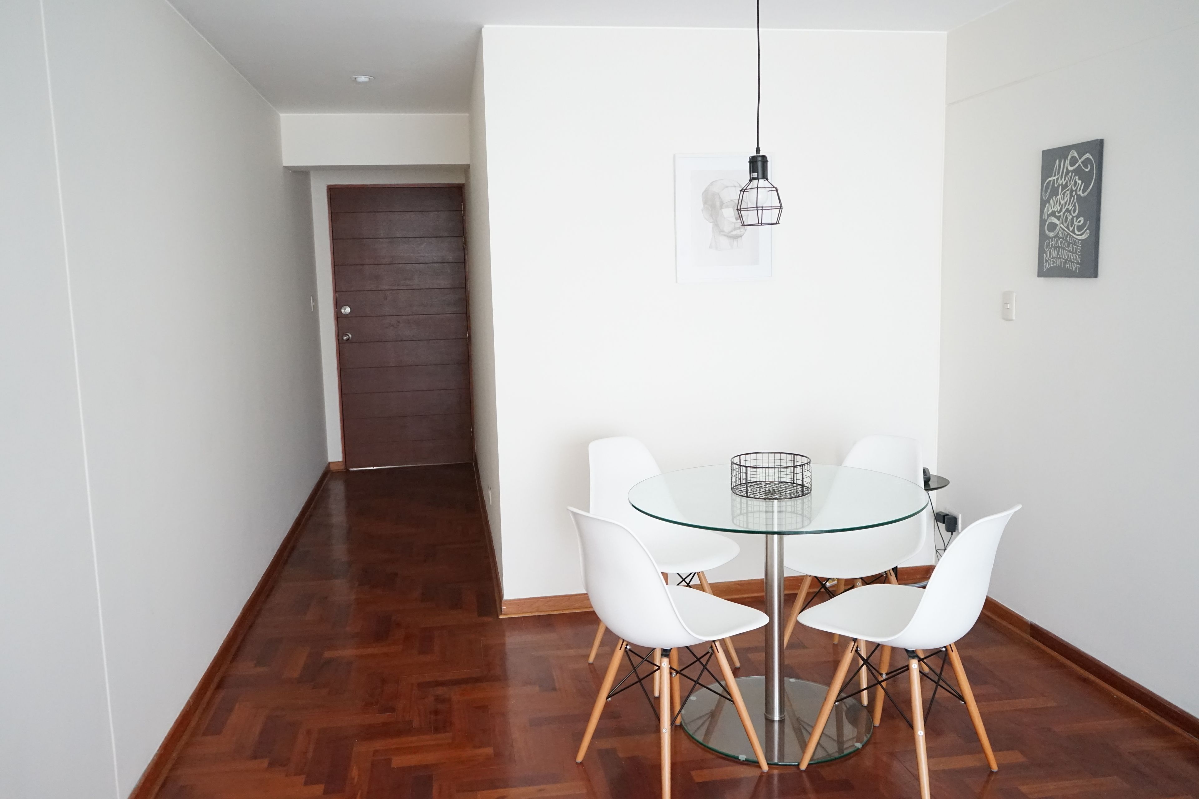 Un comedor con mesa de vidrio y sillas blancas, una puerta de madera al fondo, y un aplique en la pared.
