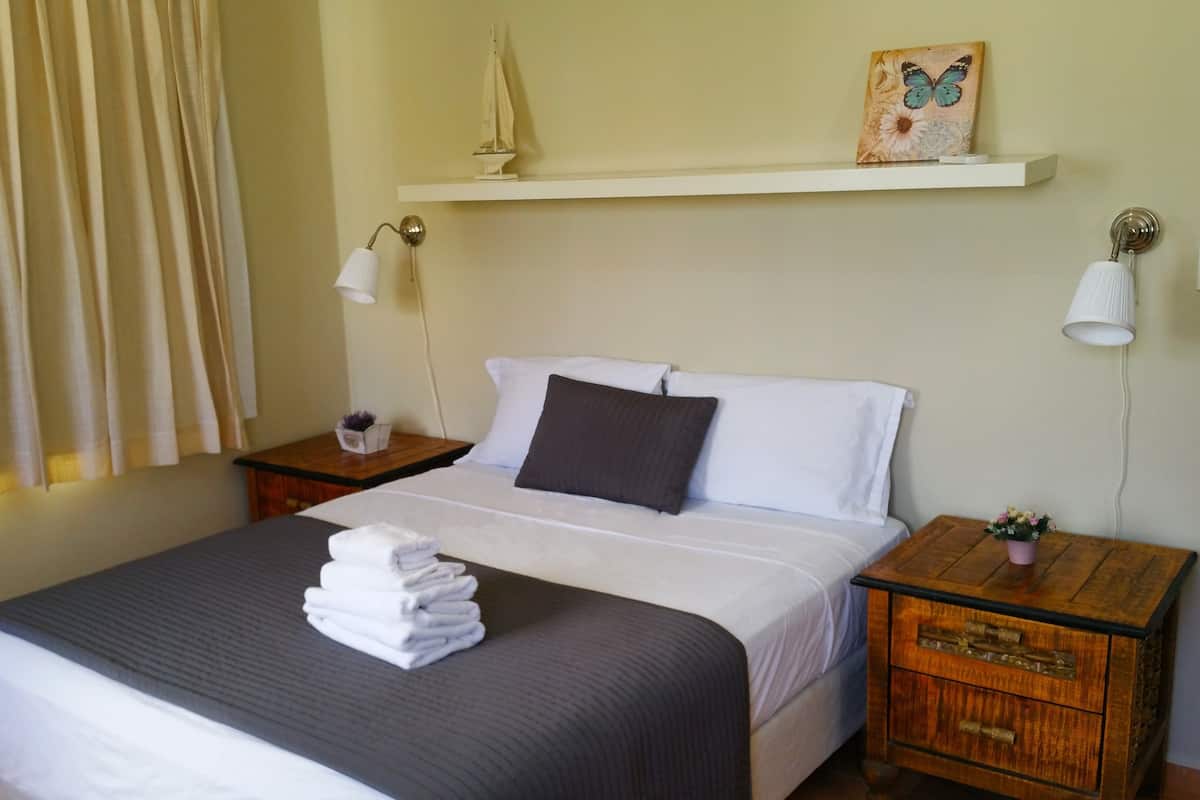 A neatly made bed with white and dark gray linens, a small nightstand with a lamp, a wooden nightstand with a plant, and a shelf with a sailboat model and a butterfly painting.