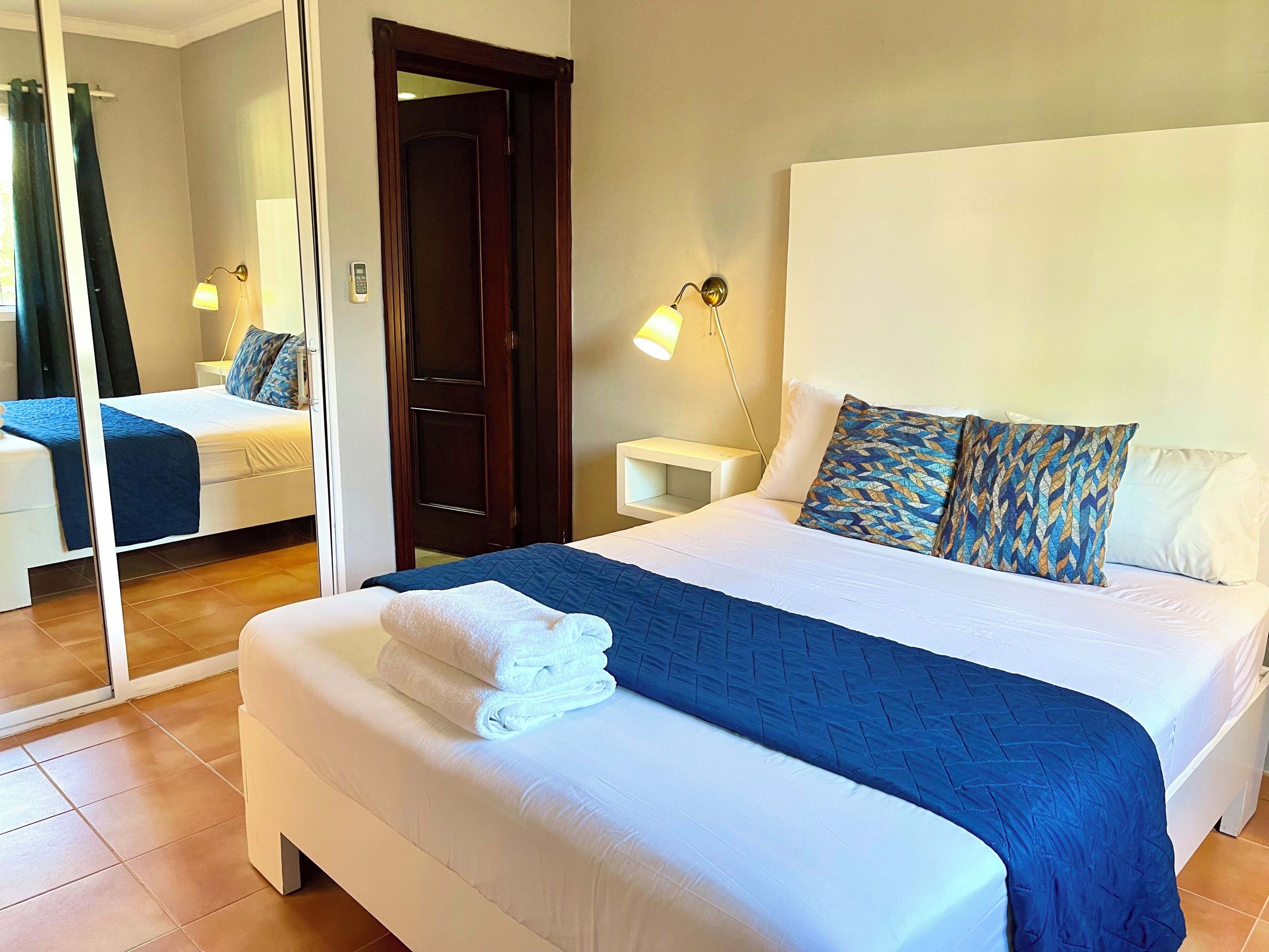 A neatly made bed with a blue quilt, white pillows, and a folded white towel. There is a bedside table with a lamp, a mirror, and a door leading to another room.