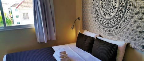 A neatly made bed with a dark quilt, a folded towel, and a wall tapestry.