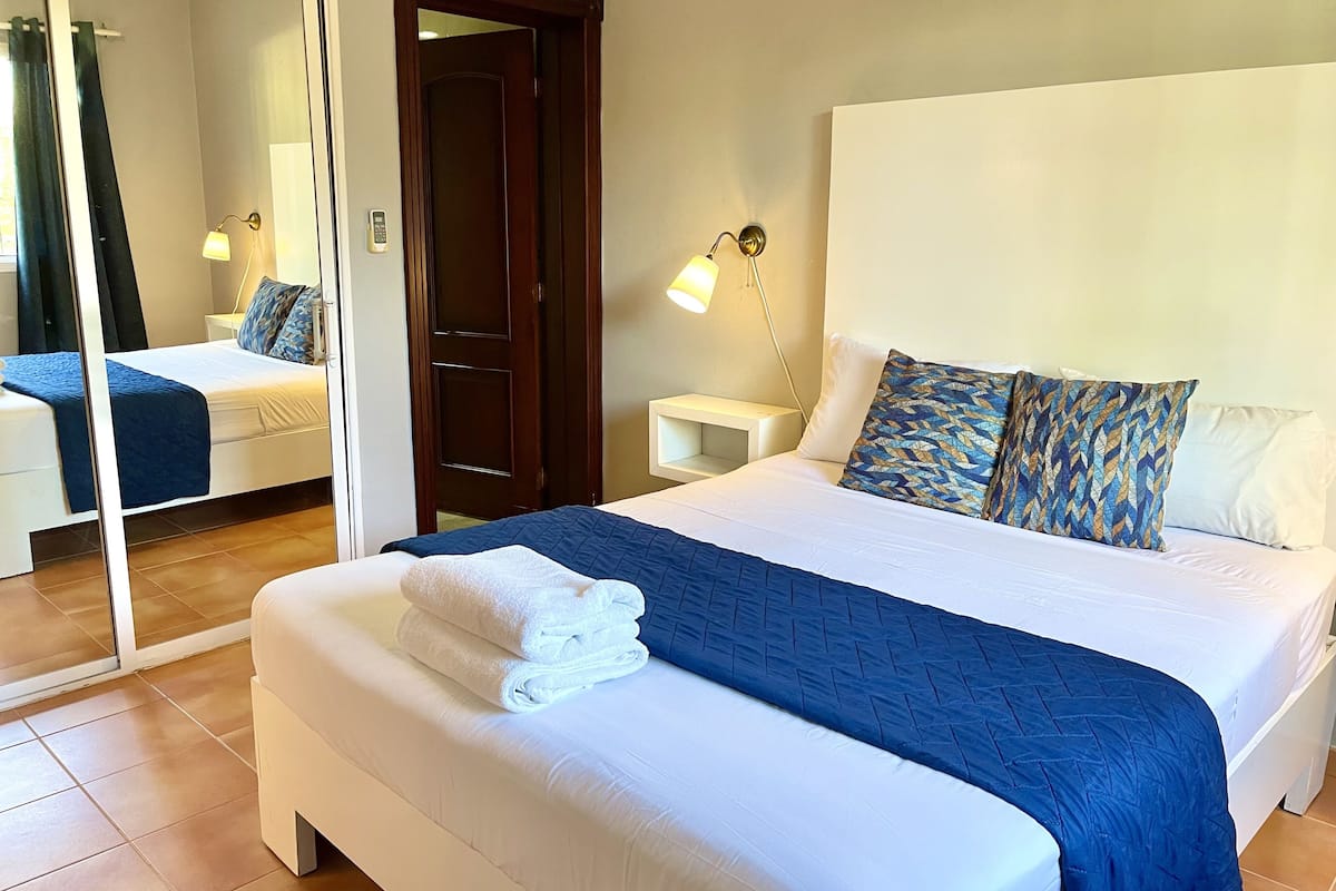 A neatly made bed with a blue quilt, white pillows, and a folded white towel. There is a bedside table with a lamp, a mirror, and a door leading to another room.