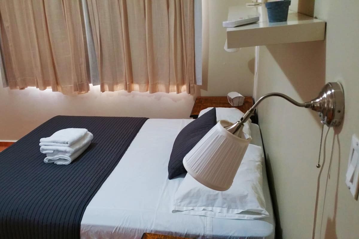 A neatly made bed with a folded towel, a bedside lamp, and a small shelf with a cup and remote control.