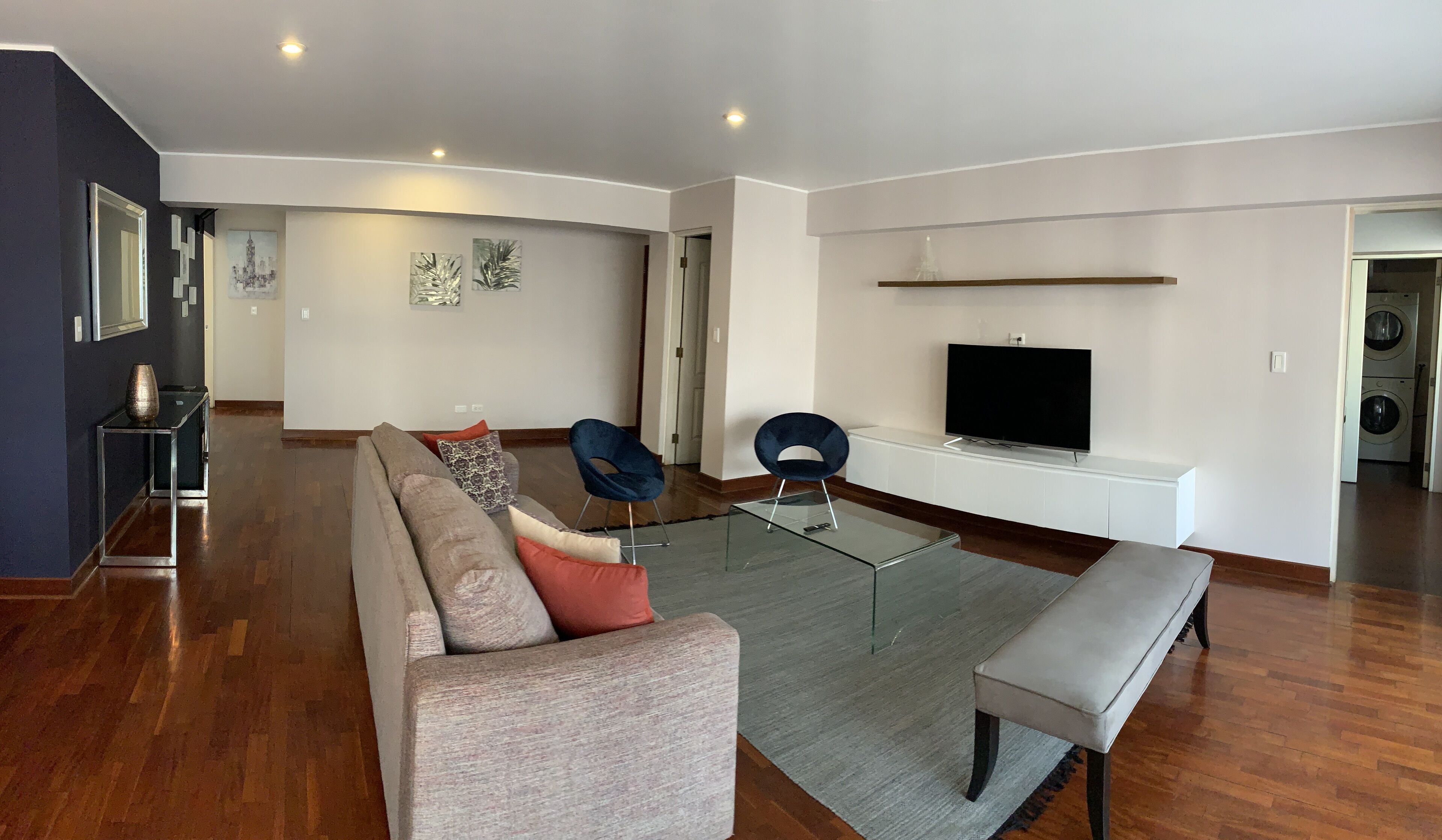 A modern living room with a sofa, two armchairs, a glass coffee table, a bench, and a flat-screen TV mounted on a white wall.