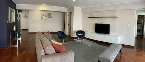 Una sala de estar moderna con un sofá, dos sillones, una mesa de centro de vidrio, un banco y un televisor de pantalla plana montado en una pared blanca.