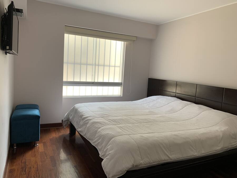 Un dormitorio con una cama grande, una ventana con persianas, un pequeño taburete azul y un televisor montado en la pared.