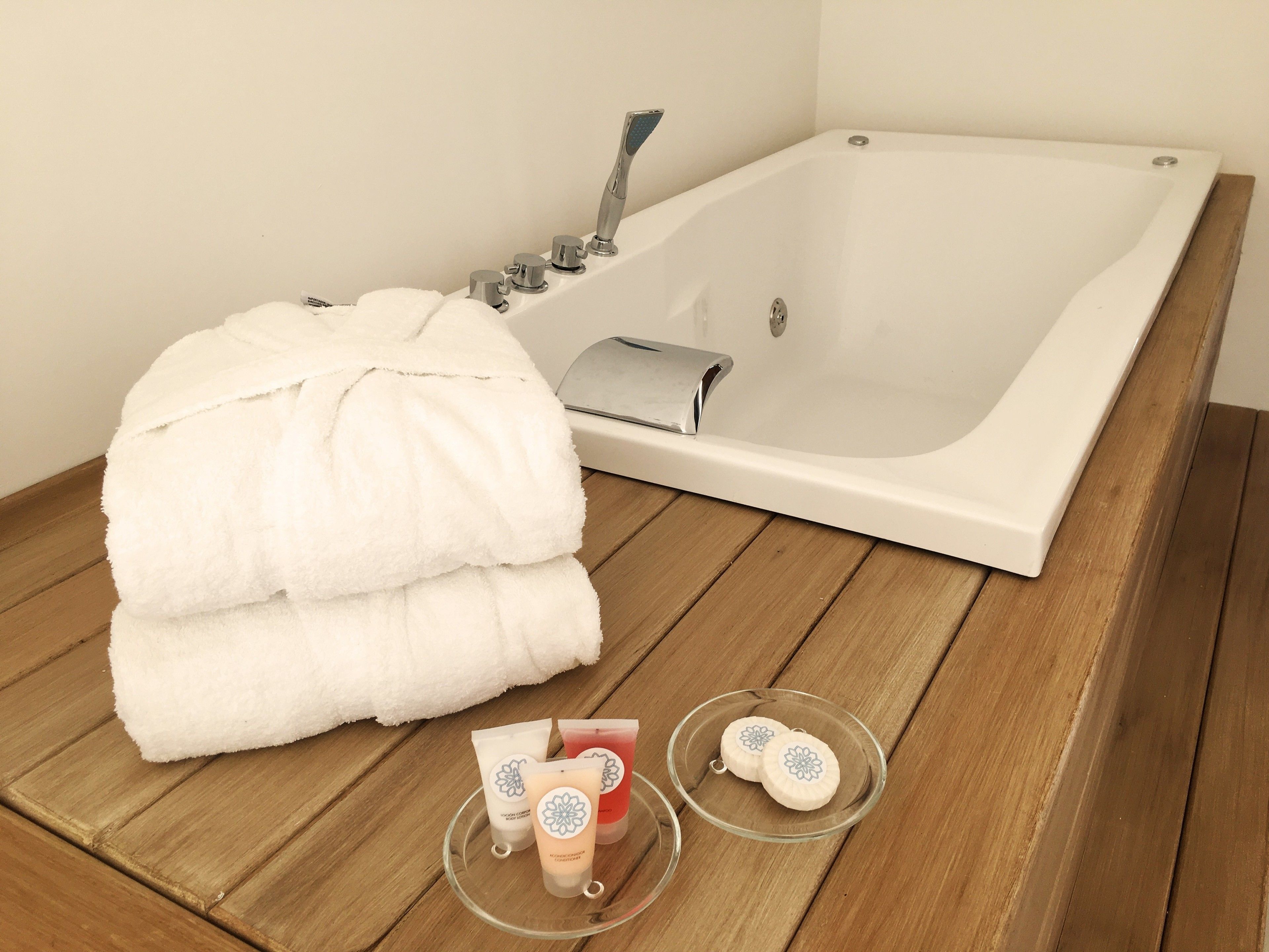 A white bathtub with a showerhead, a stack of white towels, and two glass containers with bath products on a wooden floor.